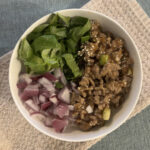 Sweet chili chicken, shredded romaine lettuce, and diced red onion on a bed of rice in a white bowl on a beige kitchen towel on a blue placemat