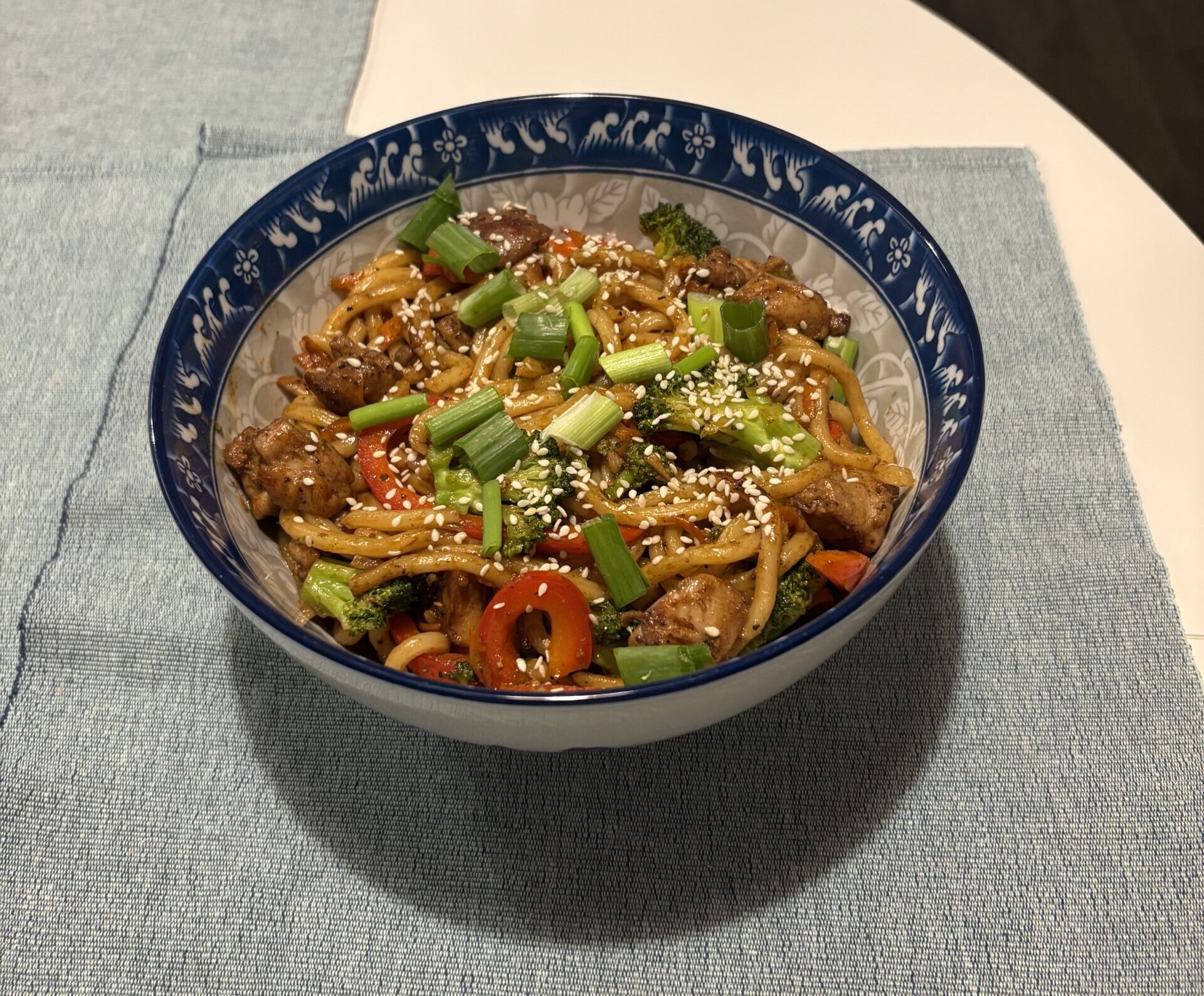 Chicken and vegetable stir-fried noodles in a blue and white bowl on a blue placemat